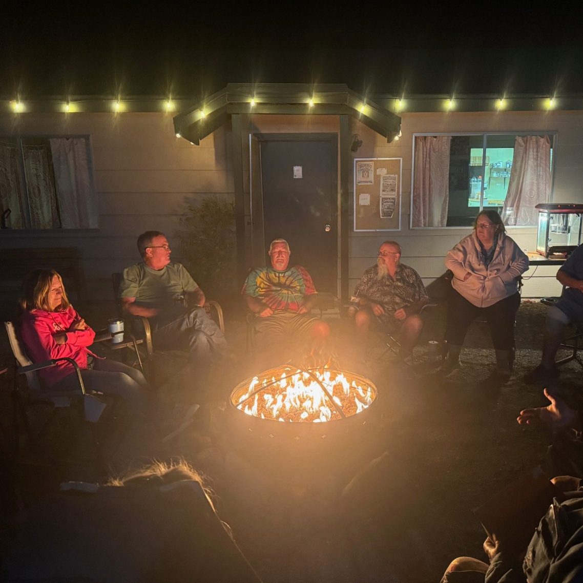 Group of people seated around a campfire at night, with warm lights in the background.