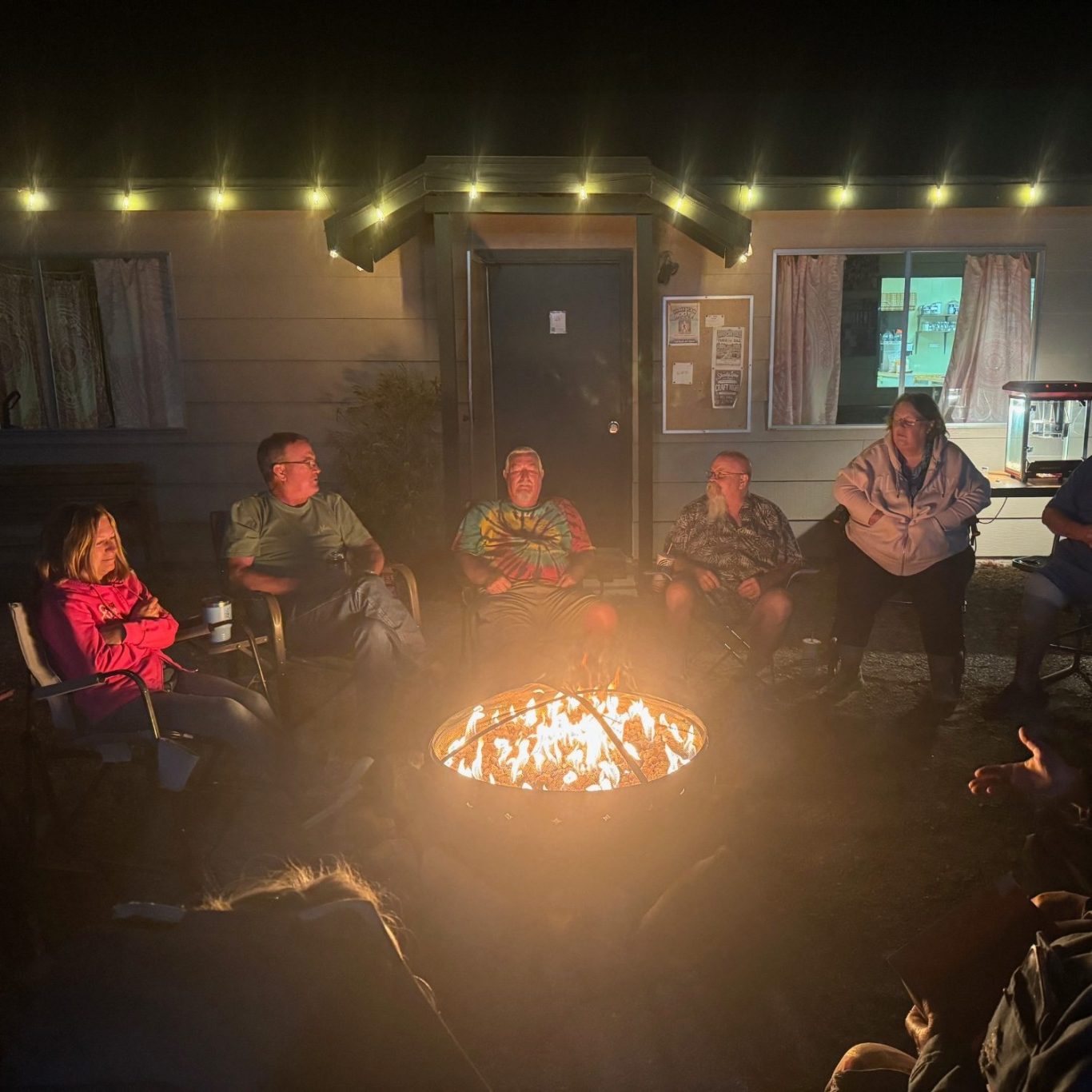 Campfire Group of people seated around a campfire at night, with warm lights in the background.